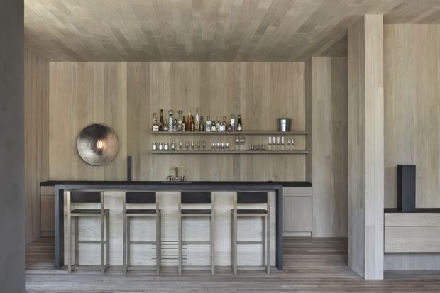 A refined built-in home bar with a long dark countertop, integrated sink, open shelving displaying bottles and glassware, and four bar stools. The space is enclosed by pale timber wall, ceiling, and cabinetry surfaces, with a substantial timber-clad structural column and a large round wall light or decorative feature.