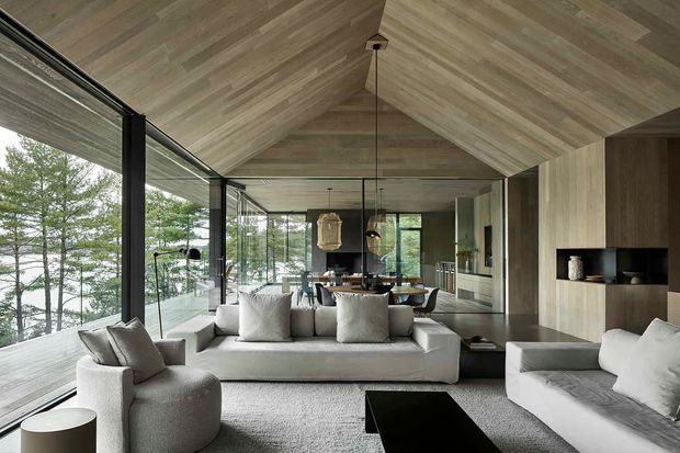 A sophisticated open-plan living and dining space with a dramatic timber-lined pitched ceiling and matching timber wall surfaces. Floor-to-ceiling glazing runs along one side, opening the room visually to a wooded landscape. The room contains a large pale sectional sofa, sculptural armchairs, a dark rectangular coffee table, a dining table with pendant lighting, and a compact kitchen or bar area beyond. The restrained palette of warm timber, soft off-white upholstery, charcoal accents, and extensive glazing creates a refined, calm contemporary interior.
