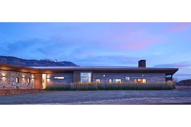 Single-storey contemporary low-profile residence photographed at dusk, with a long horizontal form and shallow roof extending over a stone-clad wing and dark timber-clad central and right sections. Warm interior and exterior lights glow through the windows, while ornamental grasses line the facade. A mountain ridge is visible in the background.