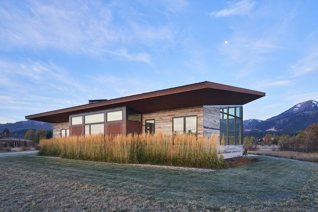 A single-storey contemporary house set in a landscaped rural or mountain setting. The compact building has a broad low-pitched overhanging roof, timber-clad walls, extensive glazing at the corner, and dark window frames. Tall ornamental grasses surround the house, with snow-dusted mountains visible in the background.