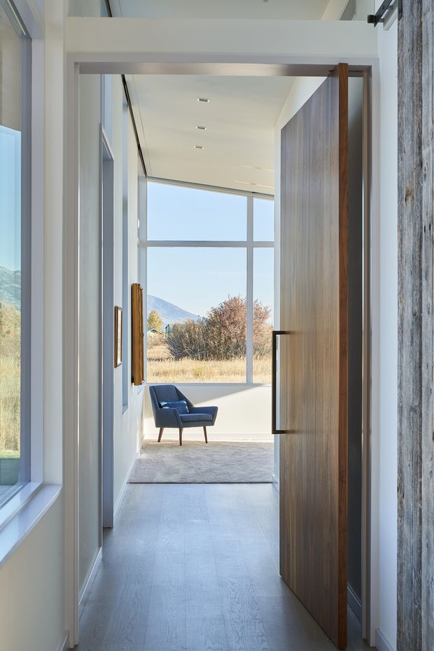 A bright modern entry hallway framed by a large timber pivot door, leading into a sunlit sitting area with a blue lounge chair. Floor-to-ceiling glazing provides expansive views of a dry landscape and mountain beyond. The space features pale timber flooring, white walls and ceiling, recessed linear lighting, and a textured stone or concrete wall surface beside the door.