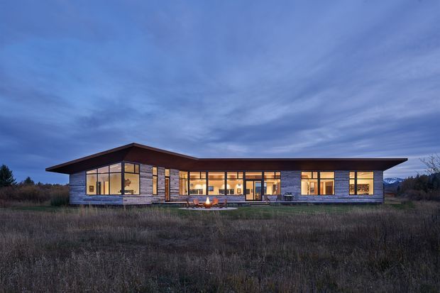 A single-storey contemporary pavilion-like house photographed at dusk, set in an open natural landscape. The long, low building has a broad shallow overhanging roof, extensive warm-lit floor-to-ceiling glazing, dark timber-toned cladding and masonry or stone piers, with a recessed central entry or terrace area.