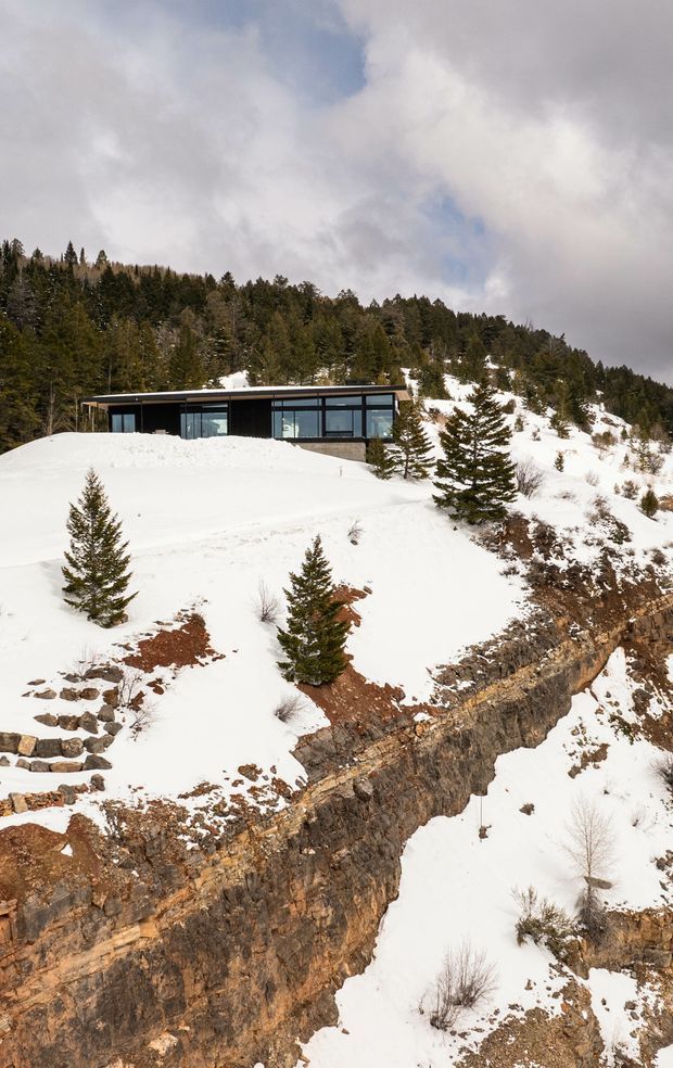 A low, modern flat-roofed house with extensive dark-framed glazing is embedded into a steep, snow-covered rocky mountainside. Dense conifer forest rises behind the building, while exposed stratified rock and sparse evergreens dominate the foreground. The image is an exterior architectural landscape photograph under a cloudy winter sky.