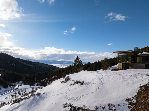 A modern dark-clad mountain house perched on a snowy, forested alpine slope. The building has a low-profile flat or gently pitched roof, extensive glazing, and a projecting upper volume, overlooking a broad snow-covered mountain valley beneath a bright blue sky.