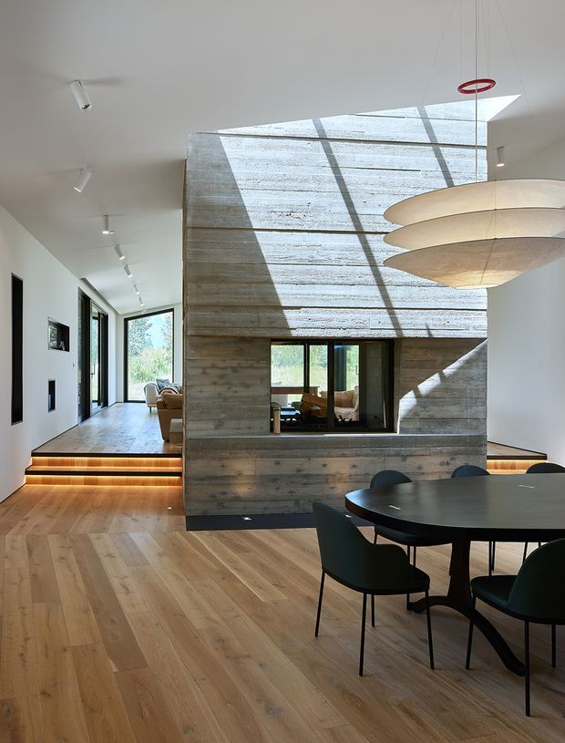 A contemporary open-plan dining and living area with a dark round dining table and upholstered chairs in the foreground. The room features a dramatic central exposed concrete block or textured concrete volume with an internal window, warm timber flooring, white walls, linear recessed and track lighting, and illuminated split-level steps leading toward a glazed living area. A large sculptural layered pendant hangs above the dining zone, while a roof skylight casts strong geometric shadows across the concrete feature wall.