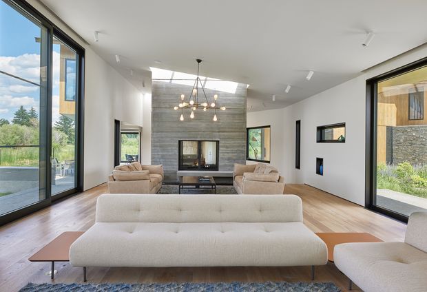Bright contemporary living room with a large cream upholstered sofa and matching seating arranged around a low coffee table. The room has wide floor-to-ceiling glazing, pale timber flooring, white walls, a dramatic textured grey stone feature wall with a fireplace and windows, and a sculptural multi-light pendant. A landscaped exterior is visible through the glazing.