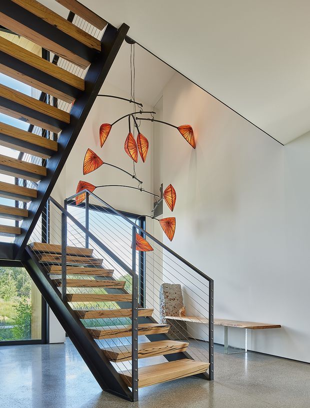 A bright modern stair hall with an open-riser timber and black metal staircase, cable railing, polished concrete floor, and a high white wall. A sculptural hanging light with orange leaf-shaped shades is suspended beside the stairs, while a simple timber bench sits along the wall. Large glazing at lower level brings in views of greenery.