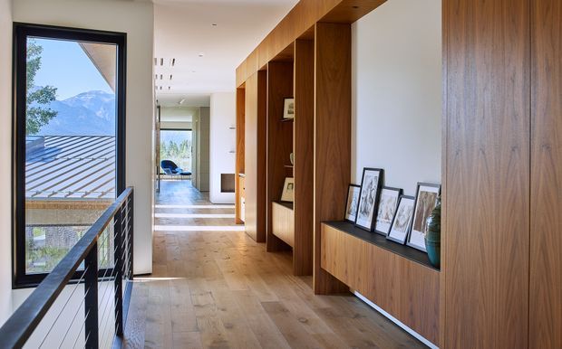 A bright contemporary hallway or entry passage with wide light timber flooring and extensive built-in timber cabinetry along one wall. The cabinetry incorporates open display niches, framed artwork, and a low console, while a black metal balustrade borders the stair opening beside tall glazing. Mountain scenery and a neighbouring roof are visible outside. The space is clean-lined, warm, and connected to a bright living area beyond.