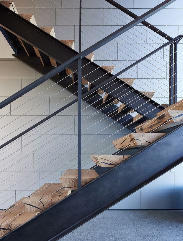 Close-up architectural view of a contemporary stairway with dark steel stringers and posts, fine cable balustrades, and sculptural natural timber treads. The stair rises diagonally through a light, geometrically tiled interior with a high glazed or open ceiling structure, creating a strong layered composition of lines and textures.