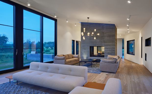 Contemporary open-plan living room with expansive floor-to-ceiling dark-framed glazing on one side, a large white sectional sofa, additional lounge seating, a low coffee table, and a dramatic grey stone-clad fireplace wall. The room has pale timber flooring, white walls and ceiling, recessed and track-style lighting, and a sculptural pendant cluster over the seating area. Green landscape is visible through the glazing.