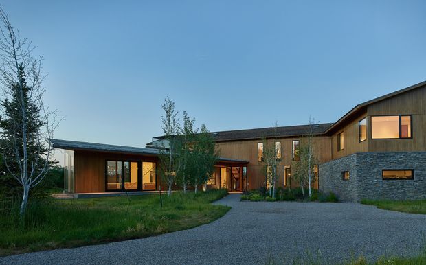 A wide exterior view of a contemporary multi-wing residence set in a landscaped site. The home combines dark timber cladding and warm timber-framed glazing with a substantial grey stone-clad lower level. Low-pitched roof forms, a recessed glazed entry, and a broad gravel driveway are visible beneath a clear blue evening sky.