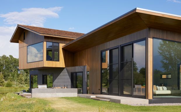 Exterior architectural photograph of a contemporary split-level home with intersecting roof forms, extensive floor-to-ceiling glazing, warm timber cladding and soffits, dark window frames, and a sheltered outdoor patio. The building sits in a landscaped suburban or rural setting with lawn and trees visible around it.