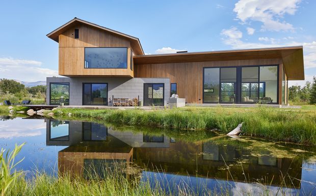 A modern single-storey residence beside a reflective pond, combining warm timber-clad volumes, dark-framed floor-to-ceiling glazing, and pale masonry walls. The building has varied pitched and low-slope roof forms, a projecting upper bedroom-like volume, and landscaped grounds with distant hills visible beyond.