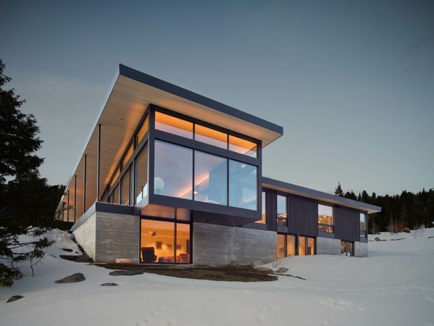 A contemporary multi-level residence set in a snowy, wooded landscape at dusk. The building has strong horizontal roof planes, extensive floor-to-ceiling glazing, dark metal-framed windows, exposed concrete walls, and warm interior lighting visible through the glass. Snow-covered ground and surrounding trees emphasize the alpine setting.