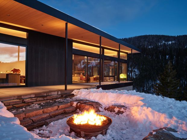 A dramatic modern mountain house exterior at dusk, set in deep snow. The low, flat-roofed structure features extensive floor-to-ceiling glazing, dark vertical wall cladding, slender dark columns, and a warm timber soffit. Stone steps lead to a snow-covered terrace with a round outdoor fire bowl, while a forested, snowy mountain slope forms the background.