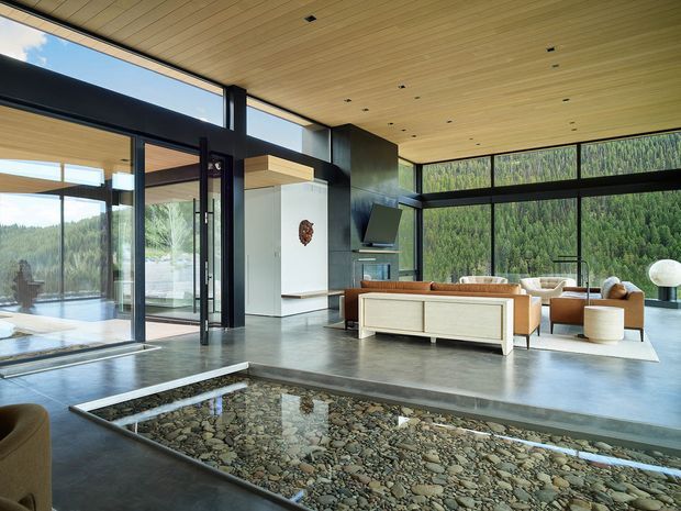 A spacious contemporary living room with expansive black-framed floor-to-ceiling glazing overlooking a forested landscape. The room features a warm timber-lined ceiling, polished concrete flooring, a sunken pebble-filled water feature or decorative void, and a minimalist sectional seating arrangement around a wall-mounted television. Open glass doors connect to an outdoor terrace or pool area.