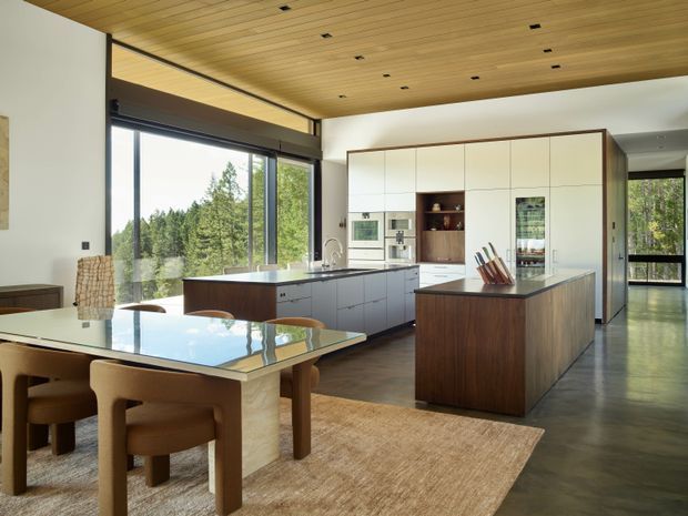 A spacious, open-plan contemporary kitchen and dining area with floor-to-ceiling glazing overlooking trees. The kitchen features a large central island with a dark timber base and stone benchtop, extensive white built-in cabinetry, integrated appliances, and a secondary peninsula. A timber dining table and upholstered chairs sit in the foreground on a natural woven rug. The room has a warm timber-plank ceiling with recessed downlights, polished concrete flooring, and a calm, light-filled atmosphere.
