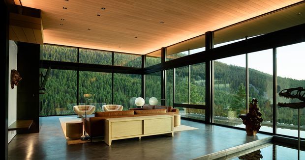 A sophisticated contemporary living room with floor-to-ceiling glazing overlooking a densely forested, mountainous landscape. The room features a low-profile seating arrangement, a large light-wood console or cabinet, dark structural window framing, polished concrete flooring, and a warm timber-lined ceiling with concealed perimeter lighting. A reflective indoor pool or water feature is visible along the foreground edge.