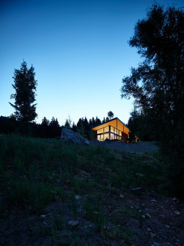 A small modern cabin-like house is illuminated at dusk in a wooded, natural setting. The building has a compact angular form with extensive floor-to-ceiling glazing, dark structural framing, and a strongly expressed pitched or gabled roof. It sits on a rocky, sloping site surrounded by tall grasses and dense trees, with a dramatic blue evening sky and warm interior lighting.
