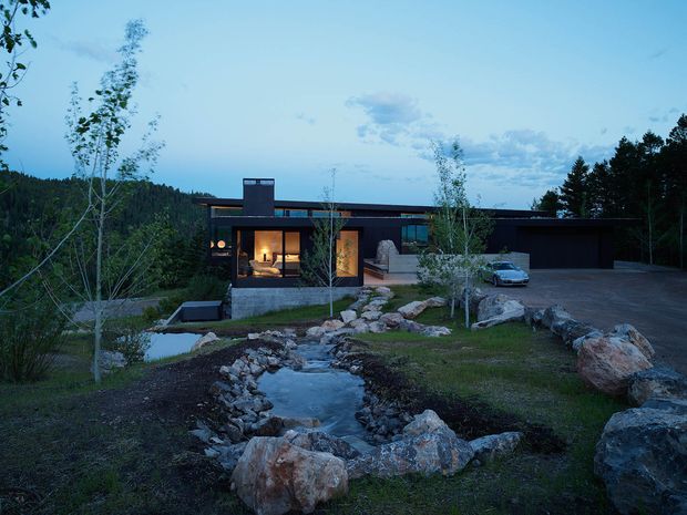A low, modern single-storey residence set within a wooded, rocky landscape at dusk. The dark horizontal facade has extensive glazing, a projecting chimney or vertical feature, and a broad flat or very low-pitched roofline. Warm interior lighting glows through the windows, while a landscaped stream or water feature lined with large natural boulders leads toward the house. A paved driveway and integrated garage are visible on the right.