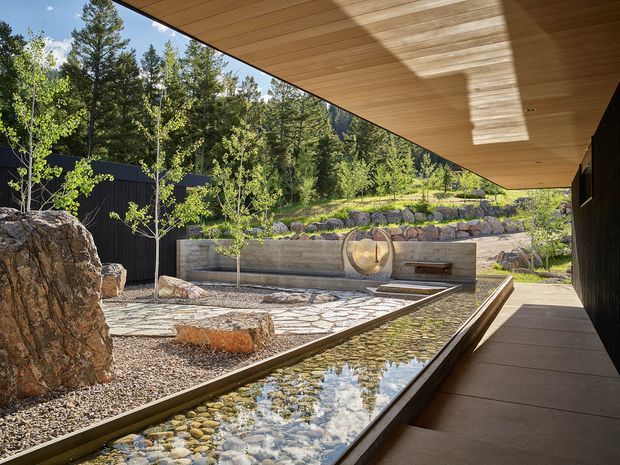A serene, landscaped courtyard beside a dark-clad building, viewed beneath a broad timber-lined roof overhang. A long, narrow reflective water feature with rounded river stones runs alongside a paved walkway. The courtyard includes sculptural boulders, young trees, layered stone retaining walls, gravel beds, and a concrete feature wall with a circular timber detail and built-in bench. The setting is natural, contemporary, and strongly integrated with a wooded landscape.