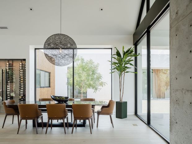 A bright contemporary dining area with a long rectangular dining table surrounded by upholstered brown chairs. Floor-to-ceiling black-framed glazing opens views toward a courtyard, while a large woven spherical pendant light hangs above the table. Pale timber flooring, white walls, a raw concrete feature wall, indoor planting, and a wall-mounted wine display create a refined modern interior.