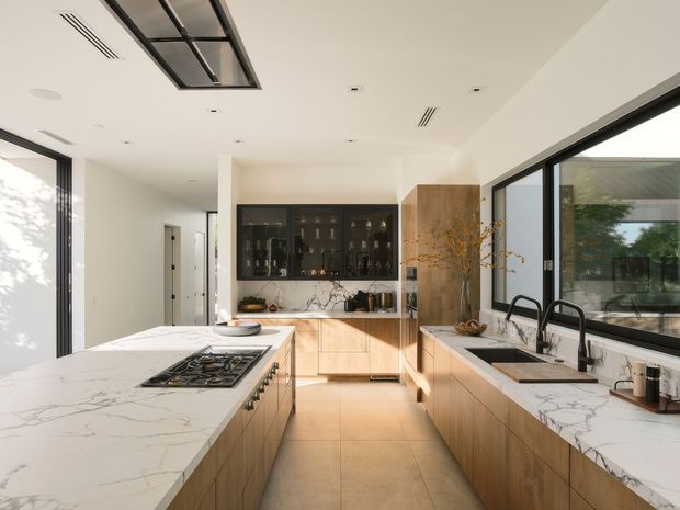 A bright, spacious contemporary kitchen viewed from the main work area. It features a large white marble-look island with integrated gas cooktop, extensive light timber cabinetry, matching stone benchtops, a dark glass-fronted drinks or display cabinet, black-framed glazing, twin black gooseneck sinks, and a roof skylight. The room has a clean, refined, warm-modern appearance with strong natural light.