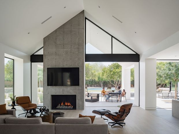 A bright, open-plan contemporary living room with a dramatic double-height pitched ceiling and expansive black-framed glazing opening onto an outdoor entertaining area. A tall raw concrete fireplace and media wall forms the central feature, while a large sectional sofa and mid-century style lounge chairs face it. Pale flooring, white walls, exposed glazing structure, and abundant garden light create an airy indoor-outdoor connection.