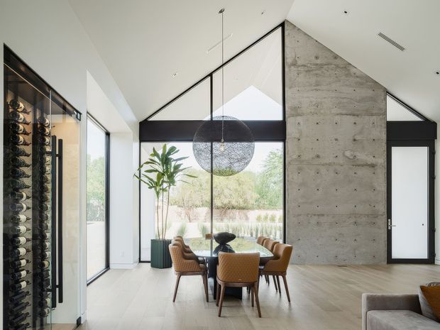 A bright, high-ceilinged contemporary dining area with a round dining table and woven upholstered chairs positioned beside expansive black-framed glazing. A large spherical pendant light hangs above the table, while a dramatic exposed concrete feature wall rises to the sloped ceiling. A glass-fronted wine storage display is visible along the left wall, and a portion of a living area appears in the foreground.