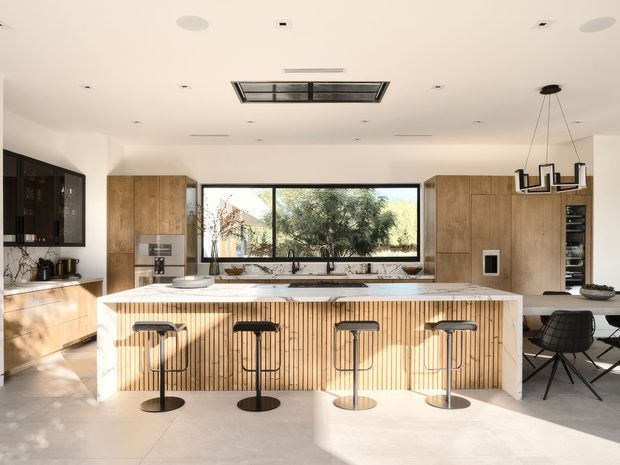 A bright, spacious contemporary kitchen viewed straight on, centred on a large island with a vertically fluted timber-clad base and pale stone benchtop. Four slim black bar stools line the island. Full-height timber cabinetry frames the rear wall, with dark cabinetry on the left, integrated appliances, a large horizontal window over the sink, and a dining area visible to the right. A linear pendant hangs above the dining area, while a large rooflight and recessed ceiling lights provide abundant illumination. The palette is warm timber, pale stone, white walls and black accents.