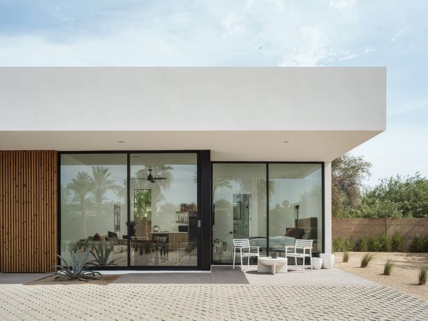 Single-storey modern home exterior with a broad flat-roofed white rendered volume, extensive dark-framed glazing, and a vertical timber screen along the facade. The paved forecourt and covered outdoor sitting area are visible, with glimpses of a contemporary living space through the glass and desert-style landscaping around the property.
