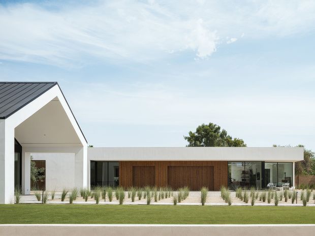 A contemporary single-storey residential home viewed from the front, composed of a prominent white gable-roofed volume on the left and a long low flat-roofed wing extending to the right. The facade combines white rendered walls, dark roof and window framing, warm vertical timber cladding around the central entry, and extensive glazing. Ornamental grasses line the paved frontage, with a broad lawn in the foreground and a bright open sky.