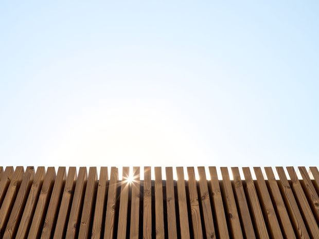Close-up upward view of a row of vertical timber slats forming a privacy screen or fence, silhouetted against a bright pale-blue sky. Sunlight shines through a narrow gap between the slats, highlighting the natural wood grain and warm brown tones.