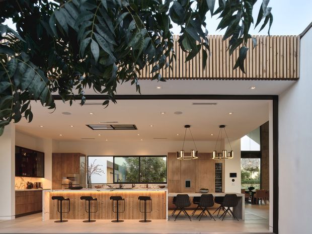 A bright contemporary open-plan kitchen and dining area viewed from an adjoining outdoor or transitional space. The kitchen features a long island with a light stone benchtop and vertically slatted timber-clad front, dark cabinetry, warm timber wall joinery, integrated appliances, and four bar stools. Behind it is a dining table with pendant lighting, while large glazed openings connect the space to greenery outside. A timber privacy screen or balcony balustrade is visible above, with a clean white ceiling and walls creating an airy, refined atmosphere.