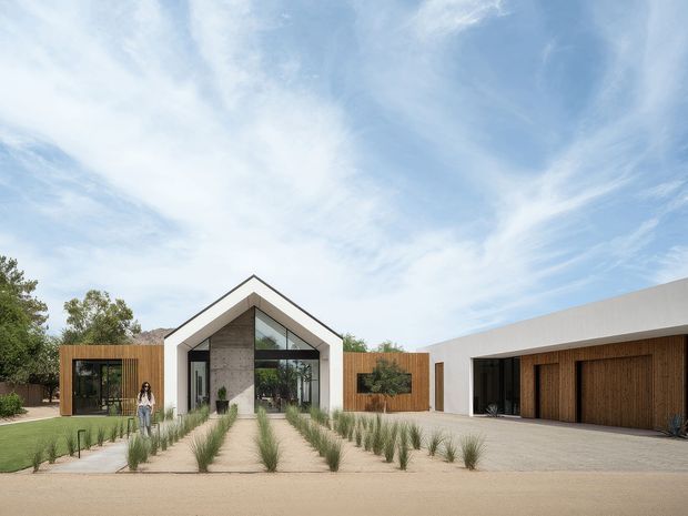 A contemporary single-storey residence viewed from the front, arranged around a broad gravel forecourt and landscaped path. The central entry pavilion has a prominent white gabled form with full-height glazing, flanked by warm timber-clad wings. A low, flat-roofed white volume on the right contains large timber garage doors. Sparse ornamental grasses, concrete stepping paths and mature trees create a restrained, desert-like landscape.
