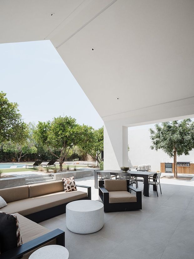 Bright, minimalist contemporary covered outdoor entertaining area with a large white geometric canopy and broad open sides. The space contains a lounge seating arrangement, dining table and chairs, pale tiled flooring, and a built-in outdoor kitchen or bar along the rear wall. A landscaped courtyard with trees and a swimming pool is visible beyond the covered terrace.