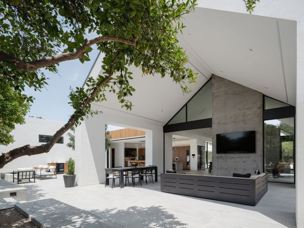 A bright, open-plan contemporary outdoor living and dining area beneath a large white flat-roof canopy. The space features a dining table, built-in dark cabinetry and entertainment unit, a large concrete feature wall with wall-mounted television, expansive black-framed glazing, and a concrete-paved terrace shaded by a mature tree. An interior kitchen and living area are visible beyond the openings.