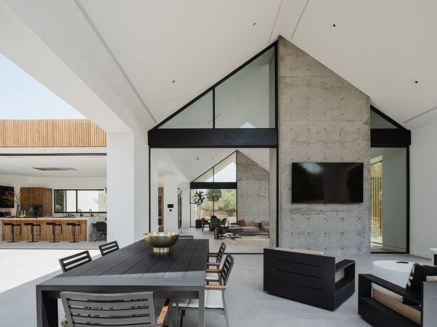Open-plan contemporary interior with a dining area in the foreground and a living room beyond, organized around a tall exposed concrete feature wall with a wall-mounted television and black-framed triangular clerestory glazing. The space has a high sloping white ceiling, polished light-grey concrete flooring, dark dining furniture, and a visible kitchen with timber cabinetry and island seating to the left. Full-height glazing and internal reflections create a bright, spacious atmosphere.