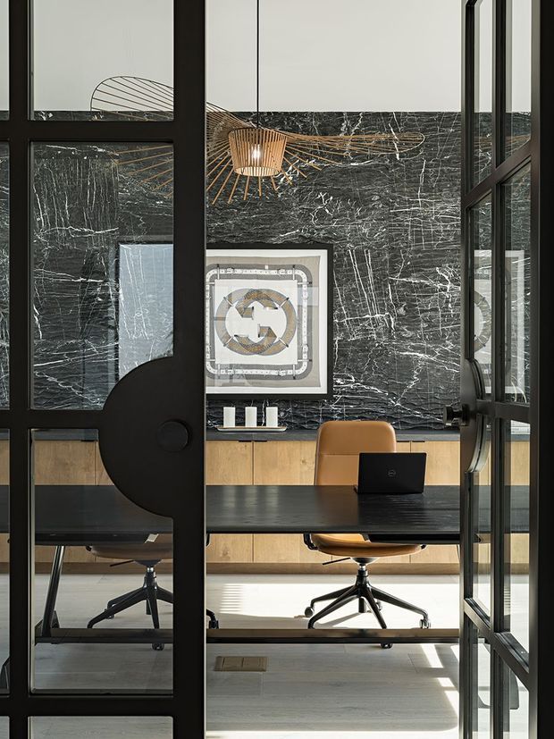 A refined home office viewed through black-framed glazed doors. The room features a dark stone-look feature wall, warm timber built-in cabinetry, a black desk, tan upholstered office chair, contemporary artwork, and a sculptural pendant light. Strong daylight enters through the glazing, creating a sophisticated, high-contrast interior.