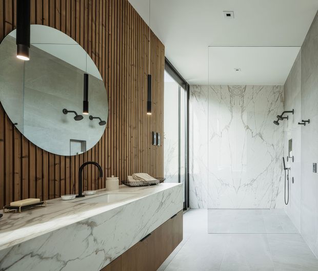 Contemporary bathroom featuring a long floating vanity with a white marble-look surface and integrated basin, warm vertical timber wall panelling, a large round mirror, dark metal tapware and pendant lights, and a spacious frameless glass shower lined in veined white stone-look slabs. The palette is light and refined with warm timber contrast.