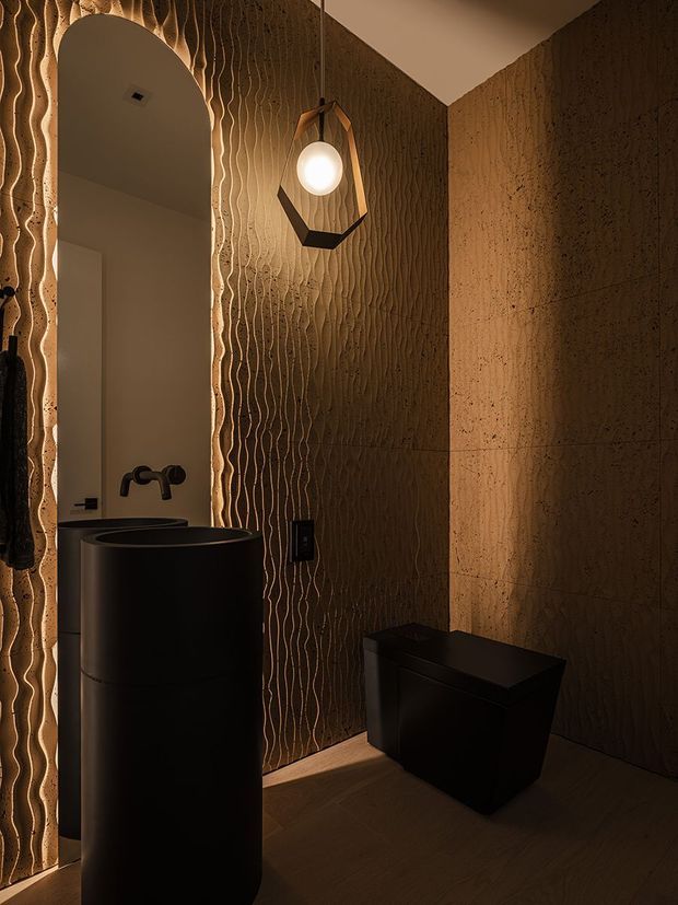 A compact, moody bathroom featuring a dramatic black pedestal basin with wall-mounted black tapware, a black toilet, and warm textured brown wall surfaces. The walls combine large stone-like panels with vertically undulating relief texture, complemented by an arched opening and a geometric pendant light with a glowing globe. The timber-toned floor and warm ambient lighting create a dark, luxurious atmosphere.