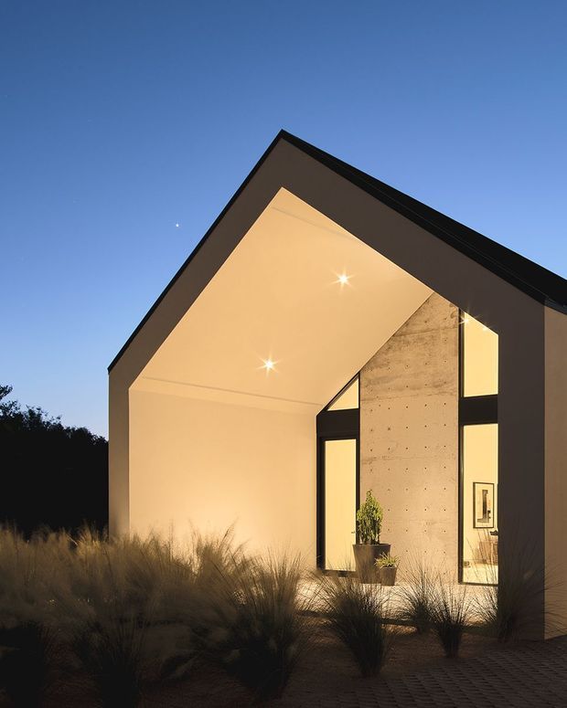 Exterior dusk view of a minimalist, single-storey gable-roofed building with a dramatic open gable facade. Dark cladding frames warm illuminated plaster soffits and large black-framed glazed openings, while a raw concrete feature wall is visible through the entry. Native ornamental grasses and textured paving occupy the foreground.