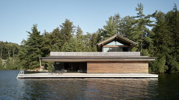 A modern two-storey lakeside house viewed from the water, with a long low rectangular form, extensive glazing, warm horizontal timber cladding, and a prominent pitched gable roof over the upper level. A narrow waterfront deck runs along the facade, with a swimming ladder at the left. Dense mature trees surround the property.
