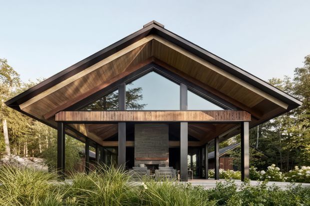 Front exterior view of a contemporary single-storey pavilion-style house set within landscaped woodland. The building has a prominent steep gable roof with broad timber-lined eaves, dark structural posts and beams, extensive glazing beneath the gable, and a projecting timber-clad balcony or entry canopy. A central dark masonry feature wall is visible through the open frontage, with ornamental planting and grasses in the foreground.