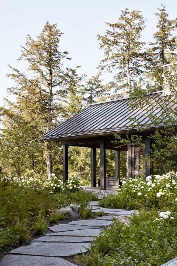A tranquil forested residential exterior viewed along a winding stone path. The building has a dark standing-seam metal gable roof, deep covered entry or porch, dark structural posts, and extensive glazing. Naturalistic landscaping with ferns, shrubs, white flowering plants, and large irregular stone pavers surrounds the structure.