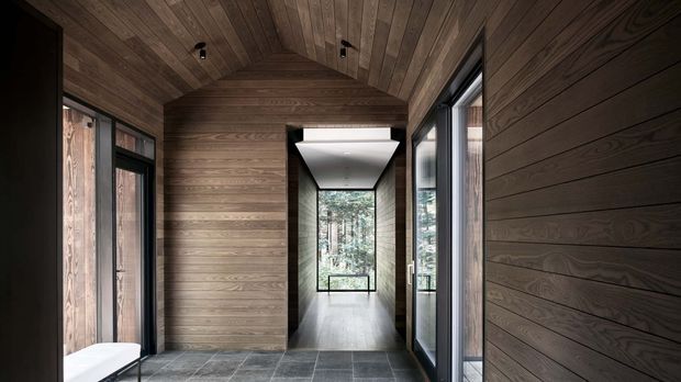 A dark, refined timber-lined interior corridor or entry passage. Horizontal wood wall panelling and a matching slatted timber ceiling frame a long view toward a bright glazed end, while large black-framed windows and doors line the sides. The floor is finished in large dark grey stone tiles, with a minimal recessed ceiling light and a glimpse of a white bench at the edge.