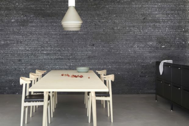 A minimalist dining room with a long pale wood dining table surrounded by light timber chairs. The room features a dark textured wall finish, smooth grey flooring, a sculptural white pendant light, and a black sideboard with a draped white textile. The restrained palette and strong contrast create a clean, refined contemporary atmosphere.
