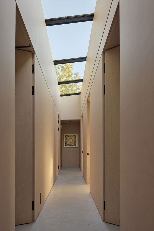 A narrow, minimalist interior hallway lined with tall flush timber-panelled doors and built-in wall surfaces. A series of overhead skylights with dark metal framing brings daylight into the corridor, with a glimpse of trees above. The pale neutral walls and smooth light floor create a calm, gallery-like passage leading to framed artwork at the far end.
