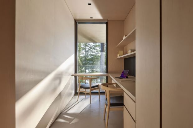 A narrow, compact home office or study with a built-in desk and open shelving along the right wall. Two timber-framed chairs sit beneath a small table by the full-height window, while warm sunlight creates strong diagonal shadows across pale walls and flooring. The space has a restrained, contemporary built-in fit-out in warm neutral tones.