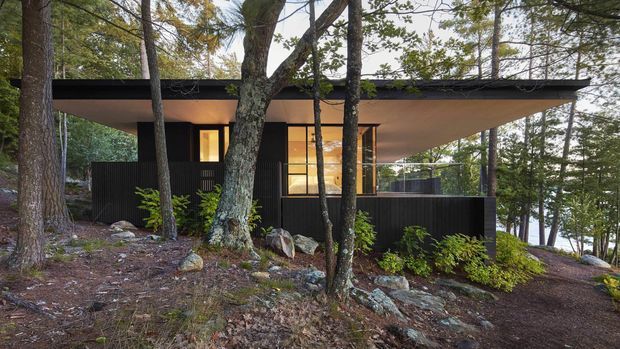A low, modern flat-roofed cabin or house nestled among mature trees on a rocky, sloped wooded site. The dark vertical-clad walls contrast with warm glowing interior glazing, while broad roof overhangs create a sheltered, horizontal composition. A glimpse of water is visible through the trees in the background.